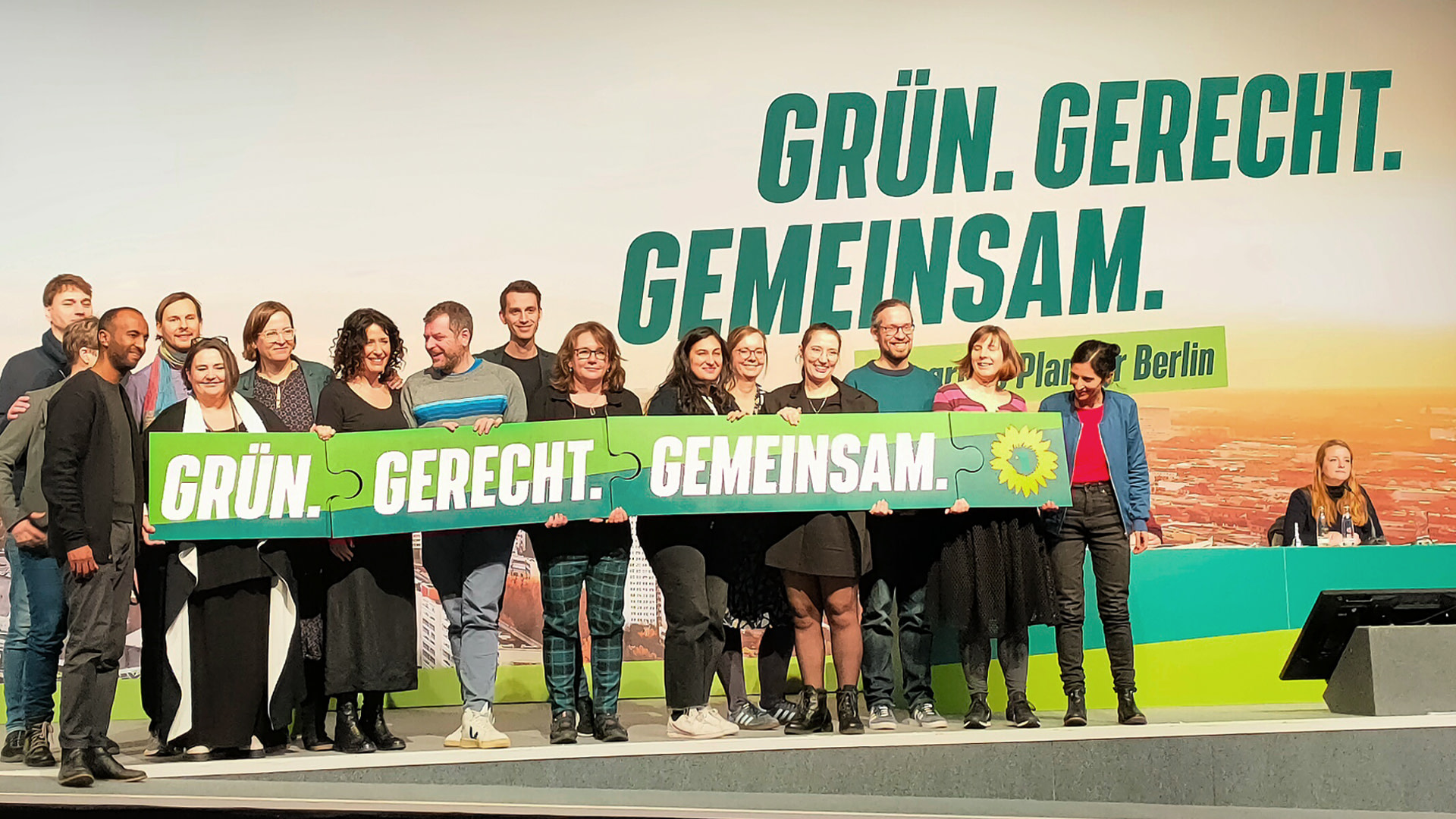 Auf dem Foto sind viele Mitglieder der Grünen Berlin zu sehen. Sie stehen vor einer Wand, worauf `Grün. Gerecht. Gemeinsam.` zu lesen ist. Dasselbe seht auch auf vier Puzzleteilen, die die Personen in die Luft halten.