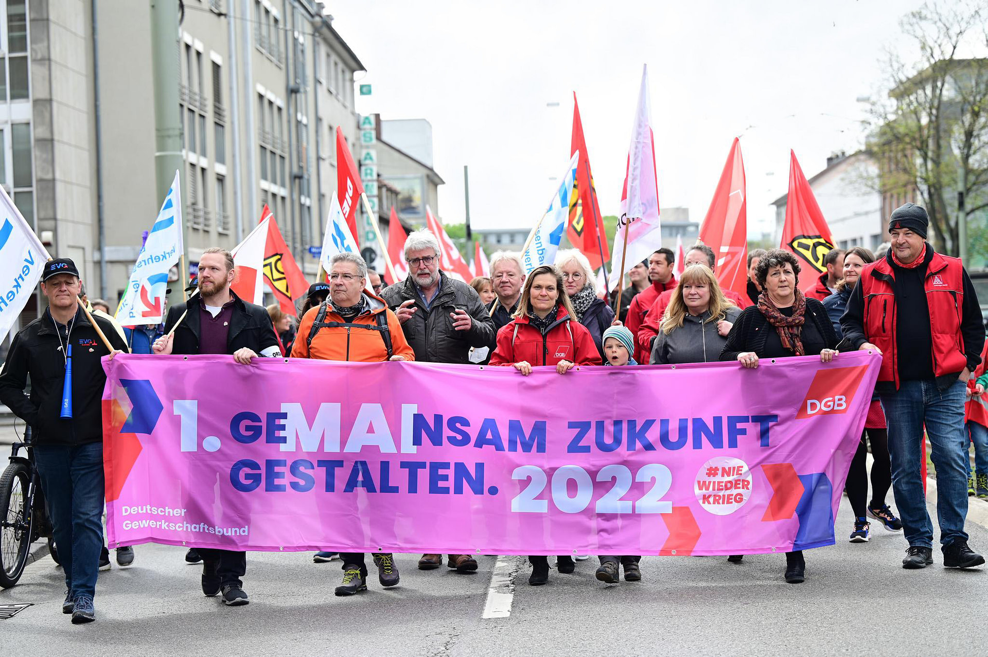 Menschen halten einen Banner zum 1. Mai, im Hintergrund wehen Demoflaggen. Auf dem Banner steht: `Gemeinsam Zukunft gestalten. - 1.Mai 2022 - #NieWiederKrieg`