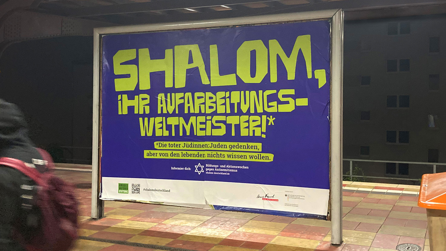 Großflächiges Plakat an einem Bahnsteig, auf lilafarbenen Hintergrund steht in giftgrün: `Shalom, ihr Aufarbeitungsweltmeister!*` und darunter: `*Die toter Jüdinnen:Juden gedenken, aber von den lebenden nichts wissen wollen.`