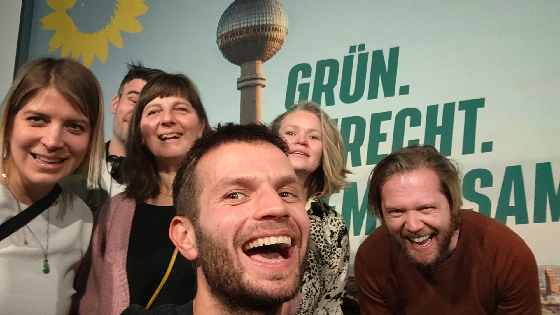 Sechs lachende Menschen aus dem Wigwam auf der Wahlparty der Grünen Berlin. Im Hintergrund ist das Backdrop zu sehen, auf welchem die Skyline von Berlin und die Headline `Grün. Gerecht. Gemeinsam.` zu sehen ist.